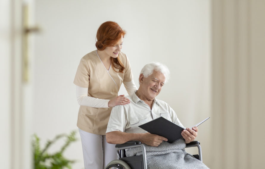 Happy elderly man in the wheelchair reading a book during visit of caregiver at home