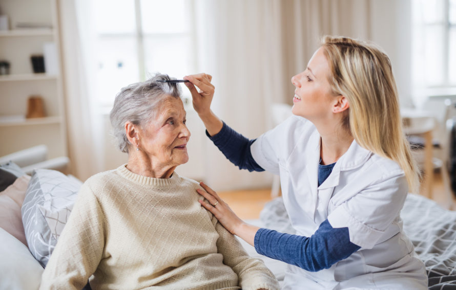 A young health visitor combing hair of senior woman sitting on a bed at home.